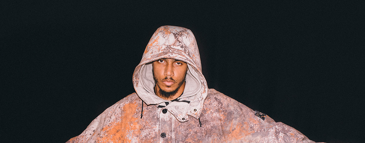AJ Tracey with his arms open wearing an orange and brown paintball Stone Island pullover jacket with the hood up