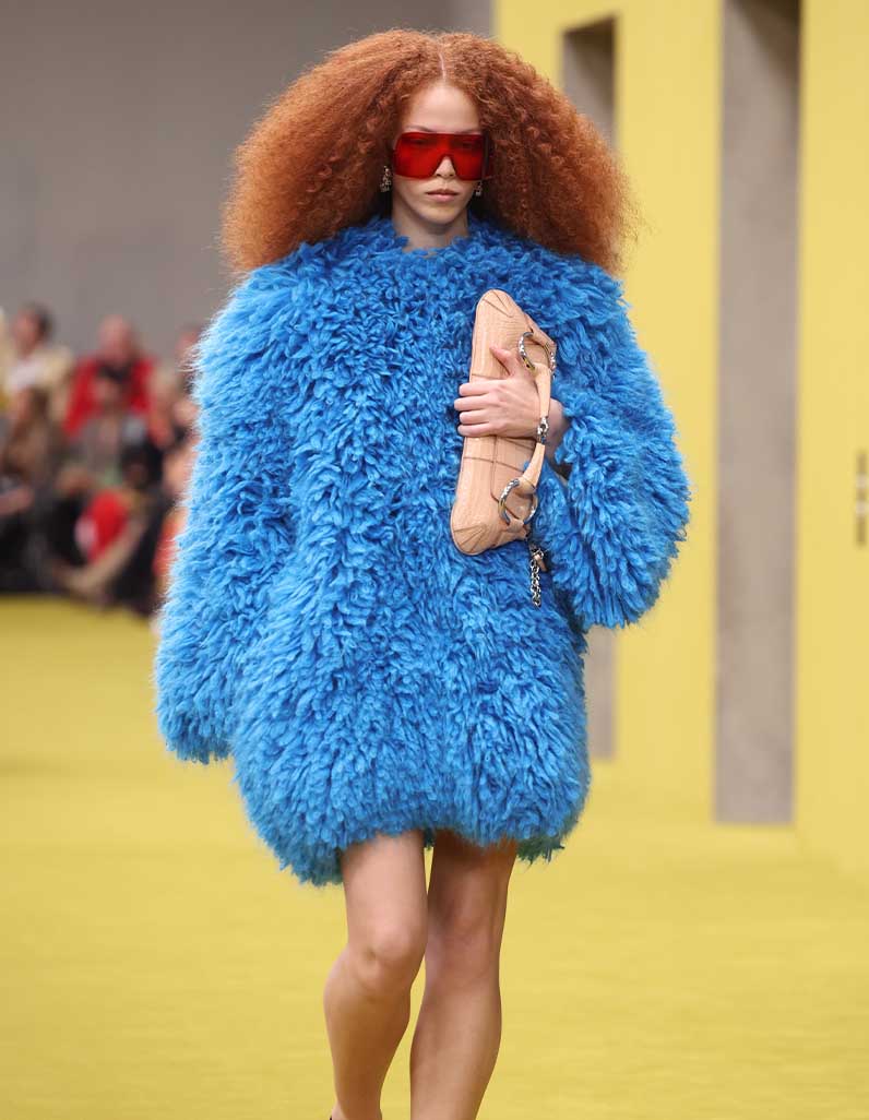 Individual wearing a bright blue over‑coat walking down a runway, against a neutral background