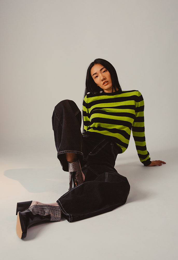 A woman with long dark hair sat on the floor, leaning back on her hands, wearing a green and black striped Ganni skit jumper, black jean Ganni flares and patchwork mock crock leather cowboy boots