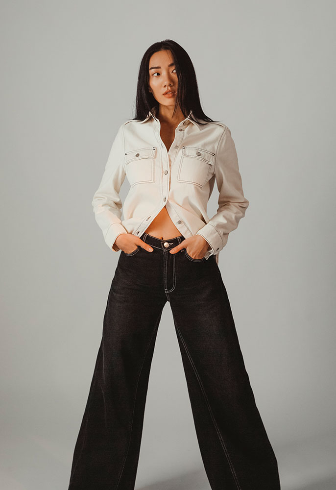 A woman stood up with her hands in her jeans pockets wearing a white denim shirt and black flared Ganni jeans with an exposed midriff